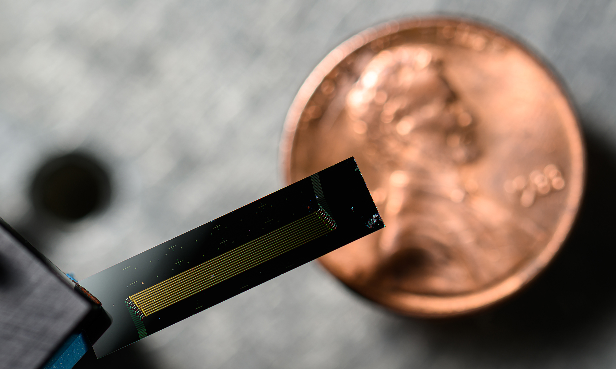 Feature image Close-up of a chip-scale laser held in front of a penny, highlighting its miniature size and intricate gold circuitry.